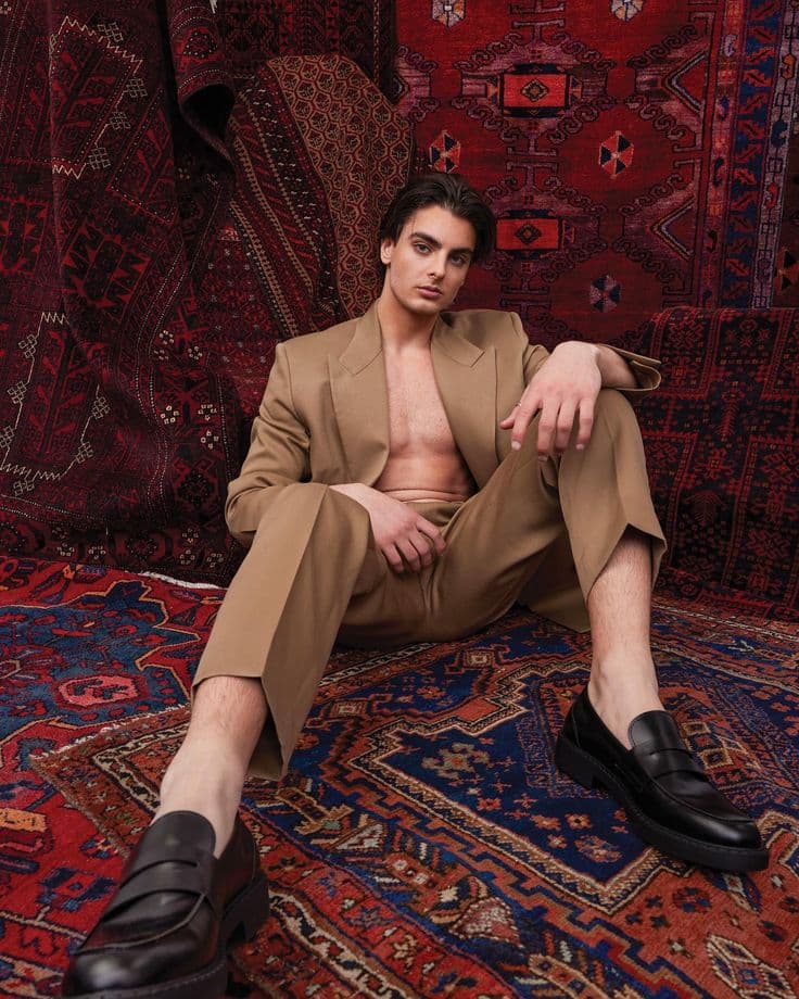 Model in a relaxed camel suit and black loafers, seated against richly patterned red and blue carpets