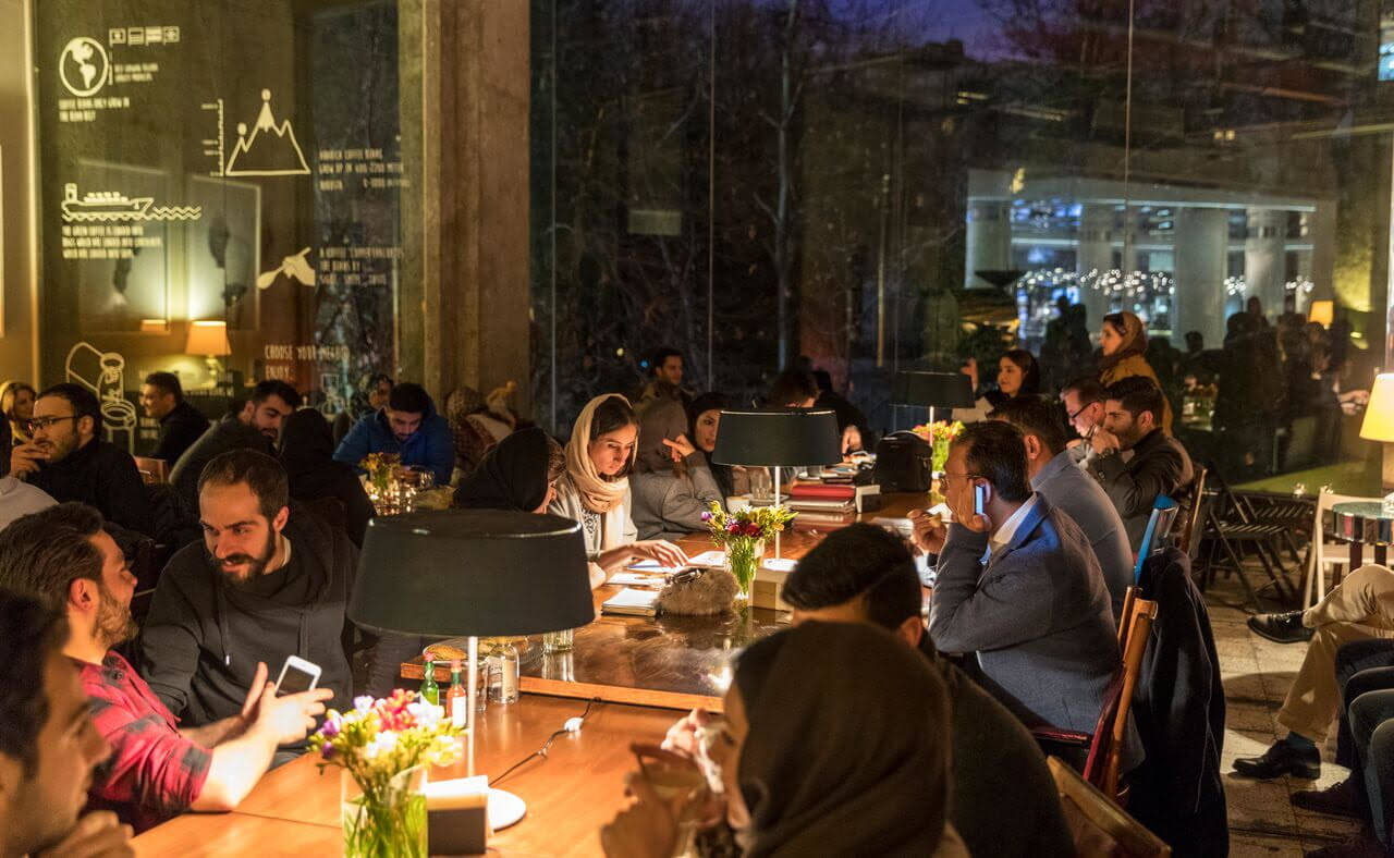 People gathering around tables in a warmly lit Tehran cafe